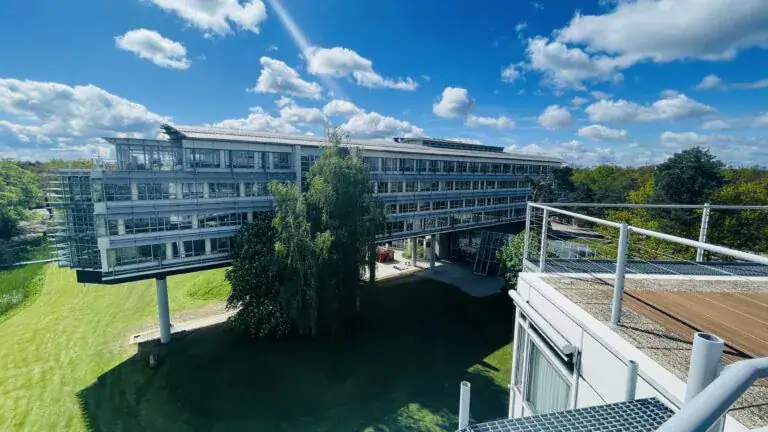 Ein modernes mehrstöckiges Gebäude mit großen Fenstern steht inmitten von grünen Rasenflächen und Bäumen unter einem blauen Himmel mit vereinzelten Wolken, aufgenommen als Aufnahmeort eines Fotos. Im Vordergrund ist ein Balkon mit einem Geländer zu sehen.